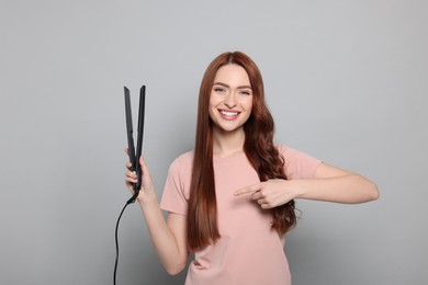 Photo of Beautiful woman pointing at hair iron on light gray background
