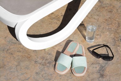 Photo of Stylish slippers, sunglasses, glass of water and grey sunbed outdoors. Beach accessories