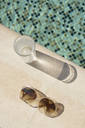 Photo of Stylish sunglasses and glass of water near outdoor swimming pool on sunny day, flat lay