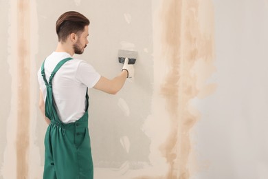 Photo of Worker in uniform plastering wall with putty knife indoors. Space for text