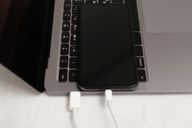 Smartphone connected with charge cable to laptop on light table, closeup. Space for text