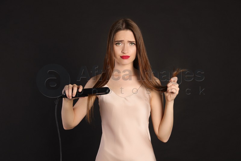 Upset young woman with flattening iron on black background. Hair damage