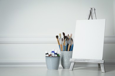 Easel with blank canvas, paints and brushes on white wooden table. Space for text