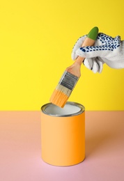 Person dipping brush into can of orange paint on pink table against yellow background, closeup. Mockup for design