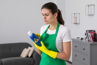 Photo of Woman looking at bottle of toxic household chemical with warning sign indoors
