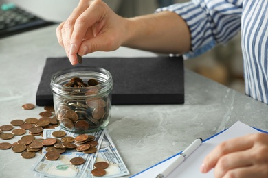 Woman putting coin into jar at grey marble table, closeup. Money savings