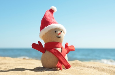Snowman made of sand with Santa hat and scarf on beach near sea. Christmas vacation