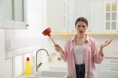 Upset young woman with plunger near clogged sink in kitchen