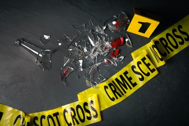 Broken bottle with blood, yellow tape and evidence marker on black slate background, flat lay. Crime scene