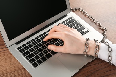 Woman with chained hand typing on laptop at wooden table, closeup. Internet addiction