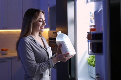 Young woman holding gallon bottle of milk near refrigerator in kitchen at night