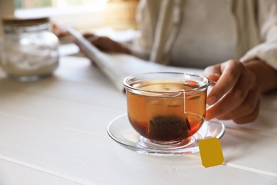 Photo of Woman holding glass cup with tea bag at white wooden table, closeup. Space for text