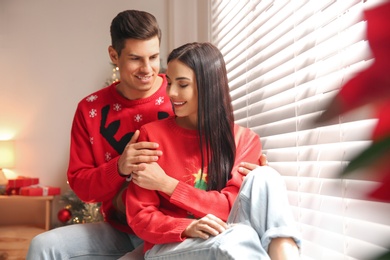 Happy couple in warm Christmas sweaters near window indoors