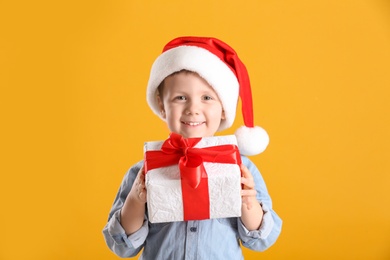 Cute little boy in Santa Claus hat holding gift box on yellow background
