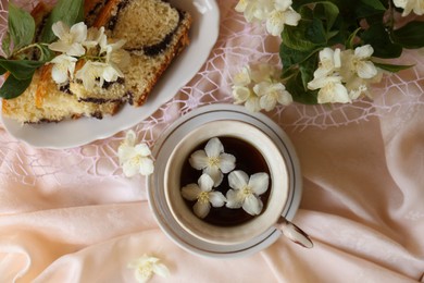Cup of aromatic tea, tasty dessert and beautiful jasmine flowers on pink fabric, flat lay