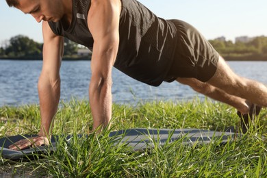 Sporty man doing straight arm plank exercise on green grass near river, closeup