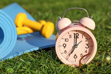 Photo of Alarm clock, dumbbells and fitness mat on green grass outdoors, closeup. Morning exercise
