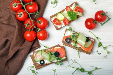 Tasty rye crispbreads with salmon, cream cheese and vegetables on white wooden table, flat lay