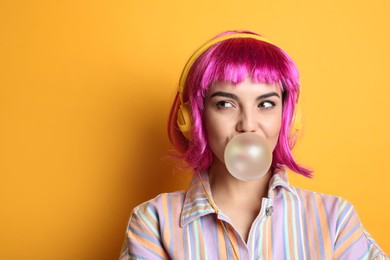 Fashionable young woman in colorful wig with headphones blowing bubblegum on yellow background, space for text