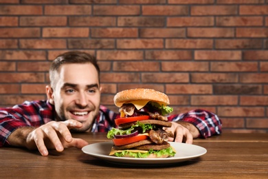 Young hungry man eating huge burger at table. Space for text