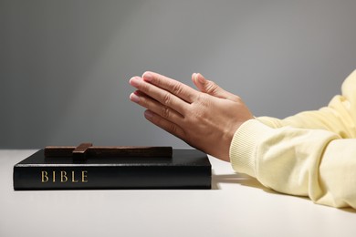 Woman praying over Bible with wooden cross at white table against grey background, closeup