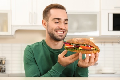 Young hungry man eating tasty sandwich in kitchen