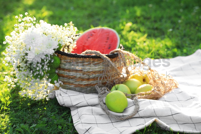 Picnic blanket with tasty fruits, beautiful flowers and basket on green grass outdoors