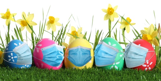 COVID-19 pandemic. Colorful Easter eggs in protective masks on green grass and flowers against white background, banner design