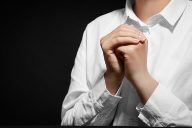 Woman holding hands clasped while praying against black background, closeup. Space for text