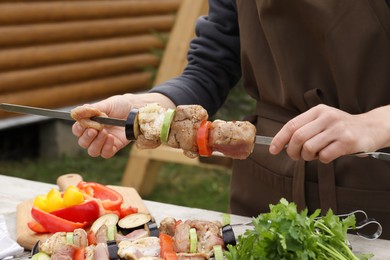 Woman stringing marinated meat and vegetables on skewer at wooden table outdoors, closeup