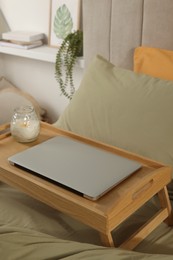Wooden tray with modern laptop and burning candle on bed indoors