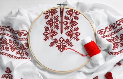 Photo of Shirt with red embroidery design in hoop, needle and thread on table, top view. National Ukrainian clothes