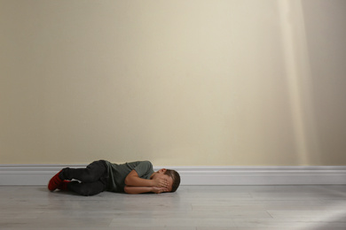 Little boy closing face with hands lying on floor near yellow wall, space for text. Child in danger