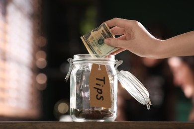 Woman putting tips into glass jar on wooden table indoors, closeup