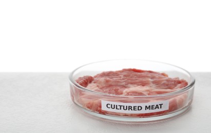 Petri dish with piece of raw cultured meat and label on table against white background
