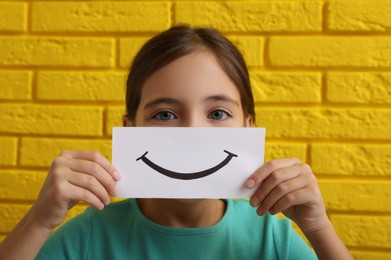 Little girl holding sheet of paper with smile against yellow brick wall
