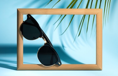 Stylish sunglasses near photo frame on light blue background