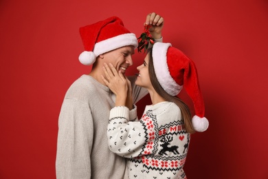 Happy couple in Santa hats standing under mistletoe bunch on red background