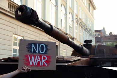 Boy holding poster with words No War near broken military tank on city street, closeup