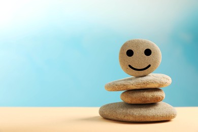 Photo of Stack of stones with drawn happy face on beige table against light blue background, space for text. Zen concept