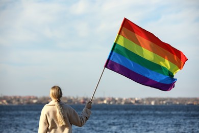 Woman holding bright LGBT flag near river, back view