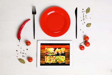 Modern tablet with open page for online food ordering, scattered vegetables, spices, plate and cutlery on white wooden table, flat lay. Concept of delivery service