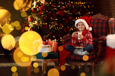 Little boy in Santa Claus cap with milk and cookies near Christmas gifts at home