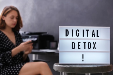 Woman using smartphone in office, focus on lightbox with phrase DIGITAL DETOX