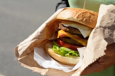 Woman holding delicious burger in paper wrap on blurred background, closeup