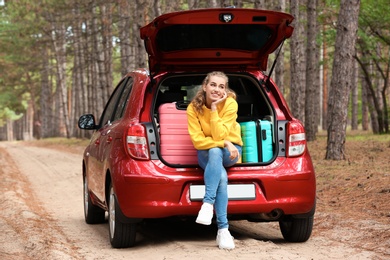 Beautiful young woman sitting in car trunk loaded with suitcases on forest road