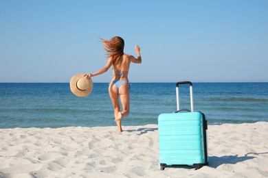 Woman in bikini running to sea and suitcase on beach, back view