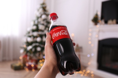 MYKOLAIV, UKRAINE - JANUARY 13, 2021: Woman holding bottle of Coca-Cola in room with Christmas tree, closeup