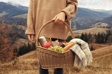 Woman holding wicker picnic basket with thermos, snacks and plaid in mountains on autumn day, closeup