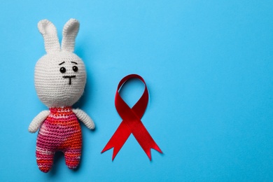 Cute knitted toy bunny and red ribbon on blue background, flat lay with space for text. AIDS disease awareness
